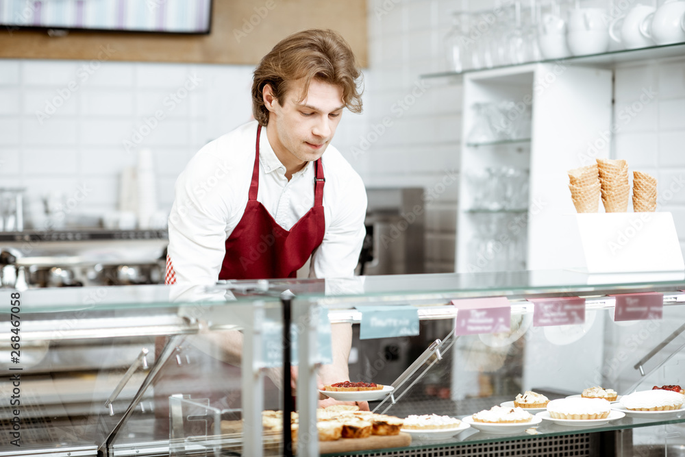 Young salesman in red apron placing sweet pastry in the refrigerator of the showcase at the modern c