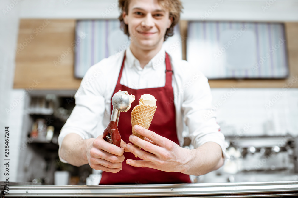 Young salesman putting ice cream ball into the waffle cone with professional tool at the pastry shop