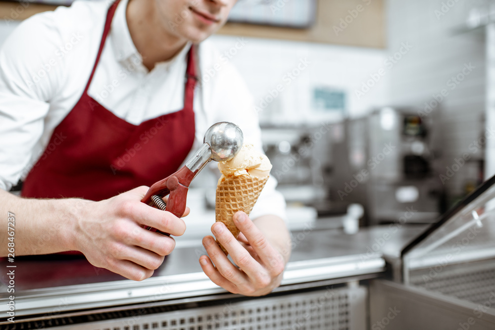 Young salesman putting ice cream ball into the waffle cone with professional tool at the pastry shop