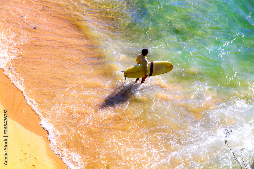 Colorful seaside with surfer walking into the sea. Surf guy carrying surfboard in bright sunny day. 