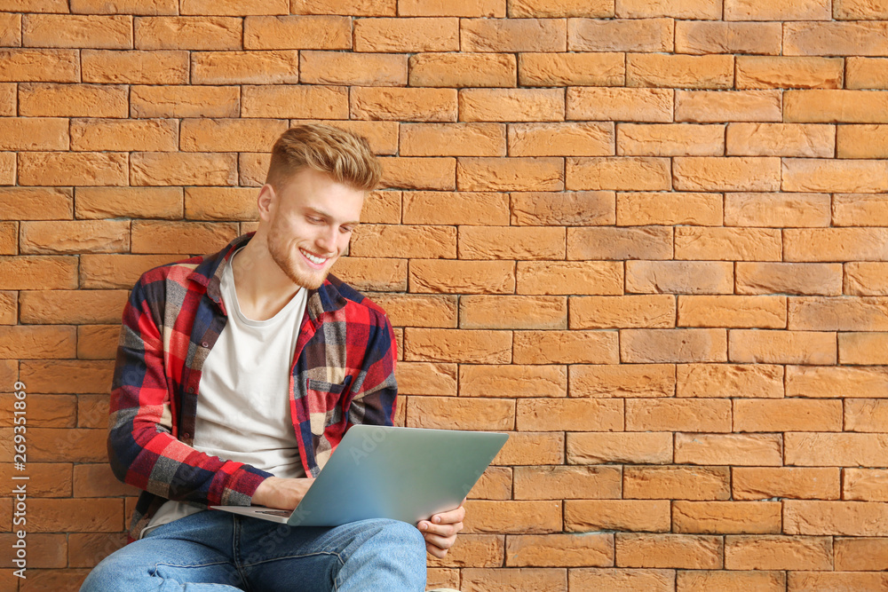 Handsome young man with laptop sitting near brick wall