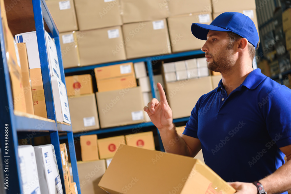 Portrait of delivery staff in blue uniform looking for particular box in warehouse