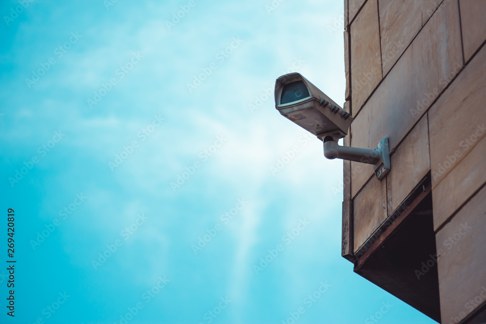 Big white surveillance camera with metallic arm placed on a building exterior with blue sky in backg