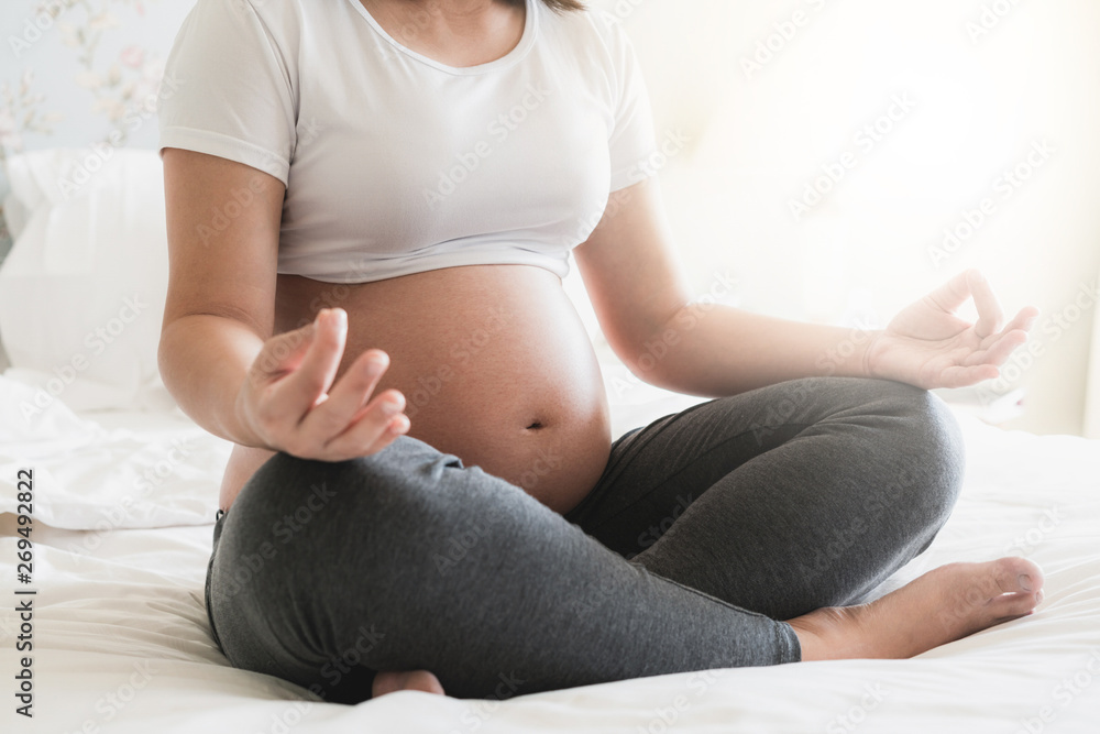 Pregnant woman doing yoga exercise on bed in bedroom at home while taking care of her child. The hap