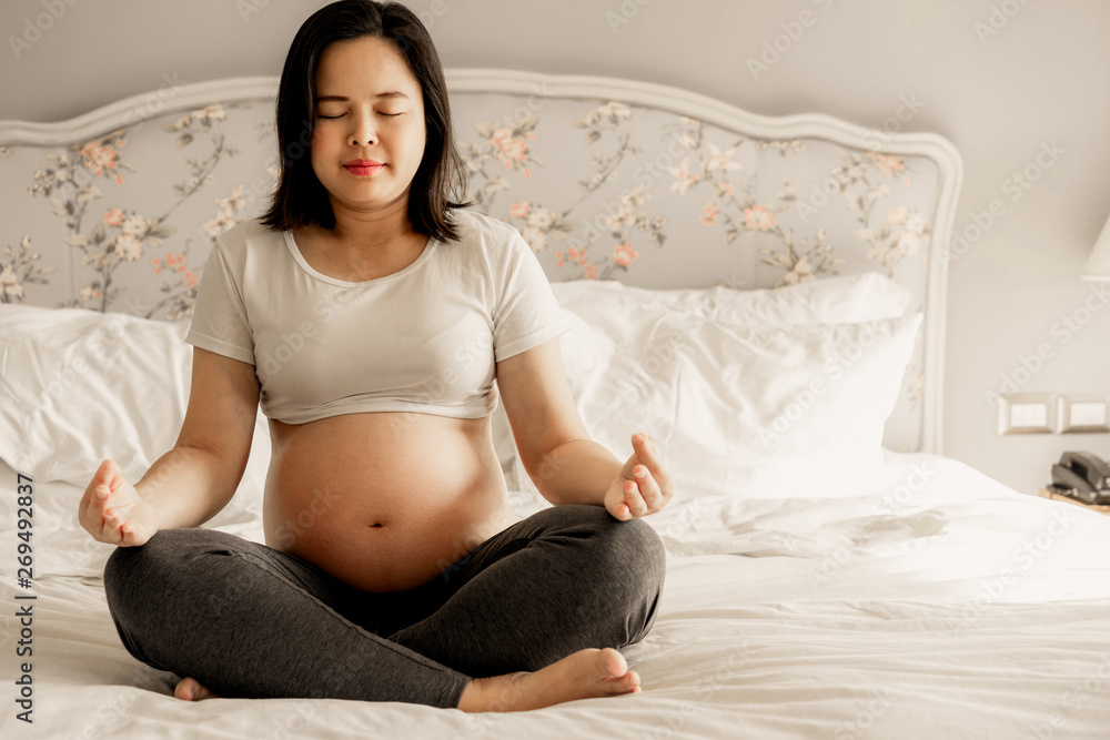 Pregnant woman doing yoga exercise on bed in bedroom at home while taking care of her child. The hap