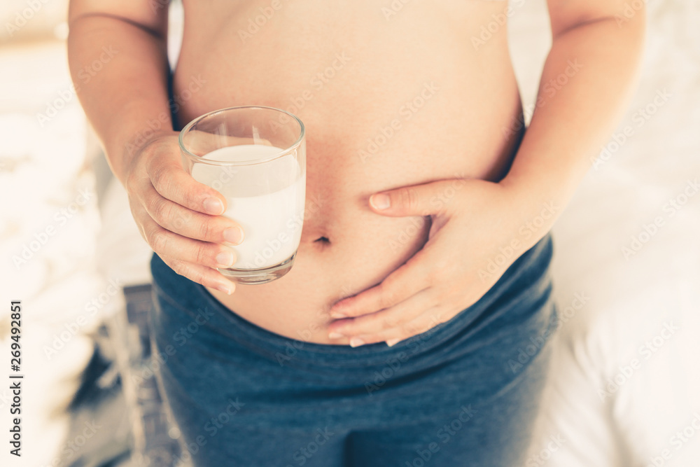 Happy pregnant woman drinks milk in glass at home while taking care of her child. The young expectin