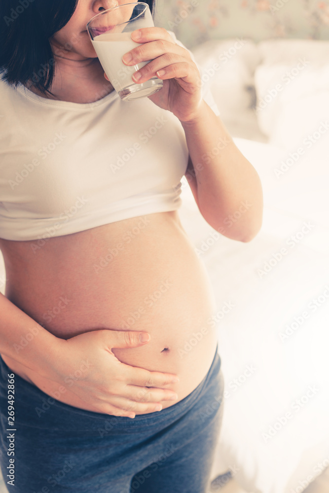 Happy pregnant woman drinks milk in glass at home while taking care of her child. The young expectin