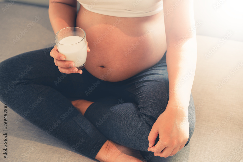 Happy pregnant woman drinks milk in glass at home while taking care of her child. The young expectin