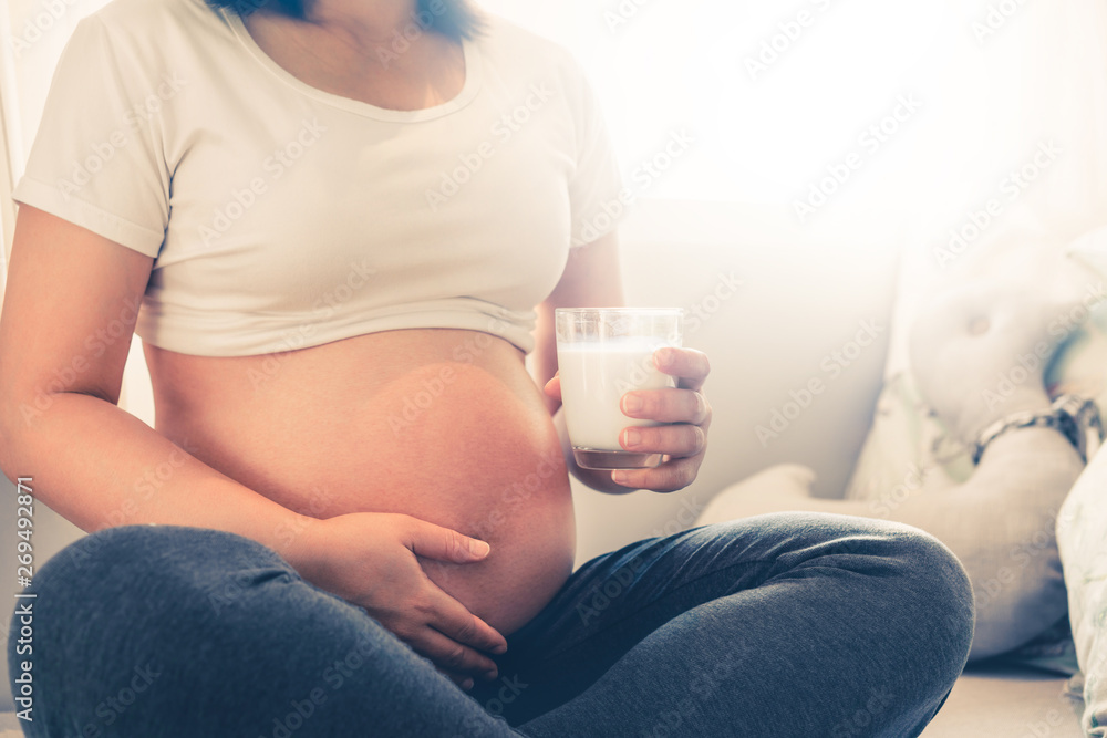 Happy pregnant woman drinks milk in glass at home while taking care of her child. The young expectin