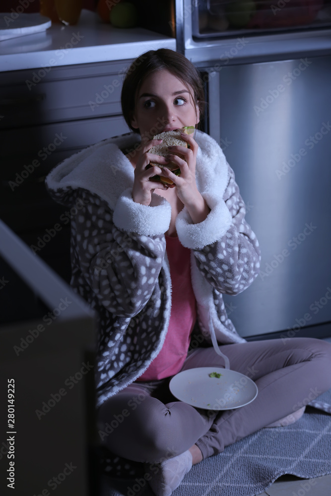 Wary young woman eating unhealthy food near fridge in kitchen at night