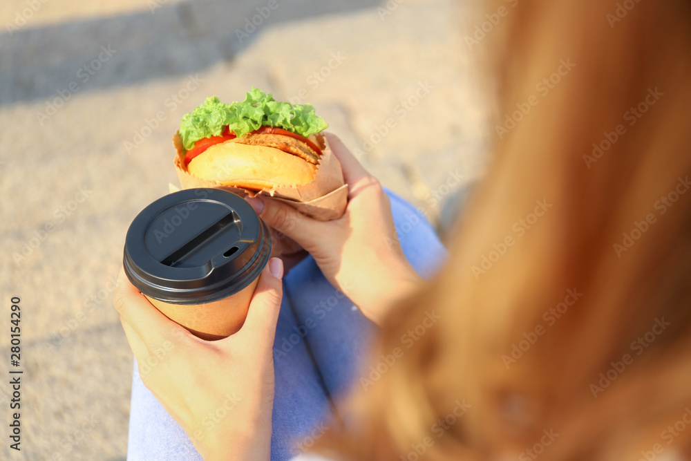 Beautiful young woman with tasty burger and cup of coffee outdoors, closeup