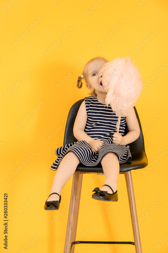 Cute little girl with cotton candy sitting on chair against color background