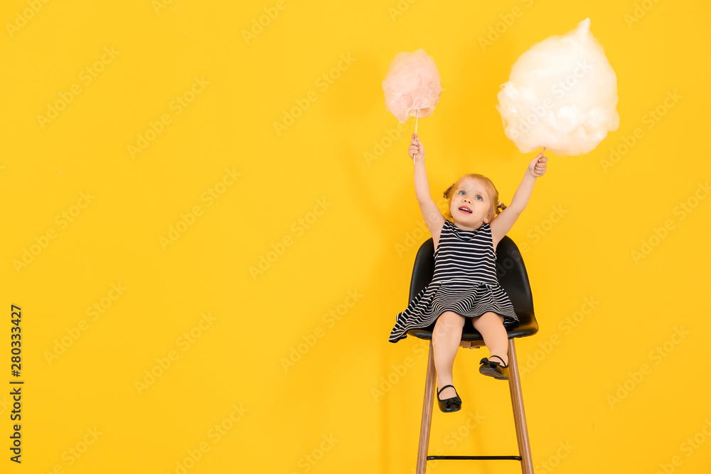 Cute little girl with cotton candy sitting on chair against color background