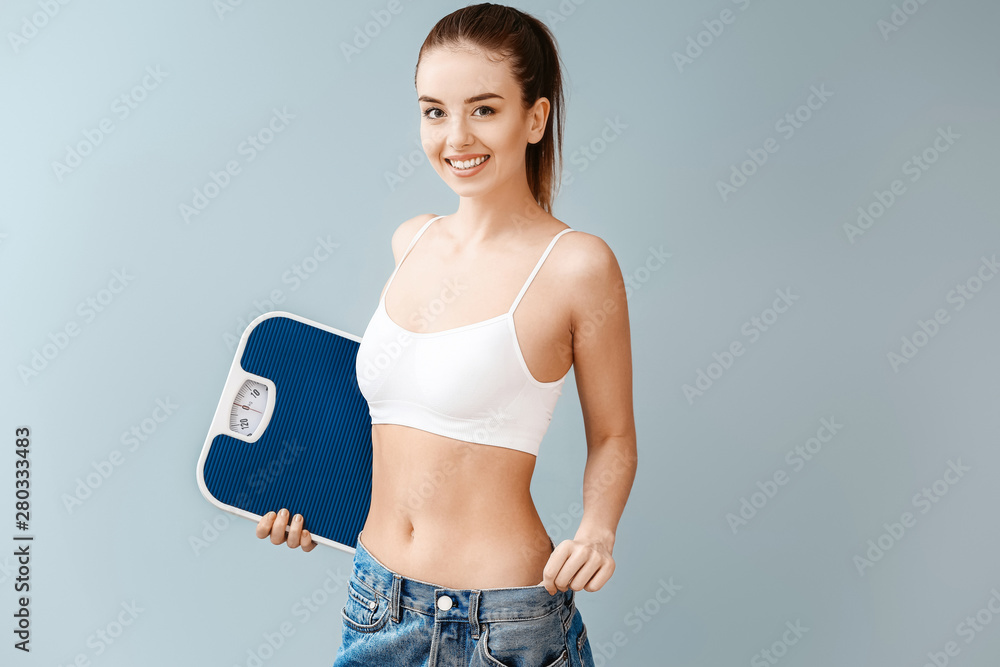 Young woman in loose jeans and with scales on color background. Weight loss concept