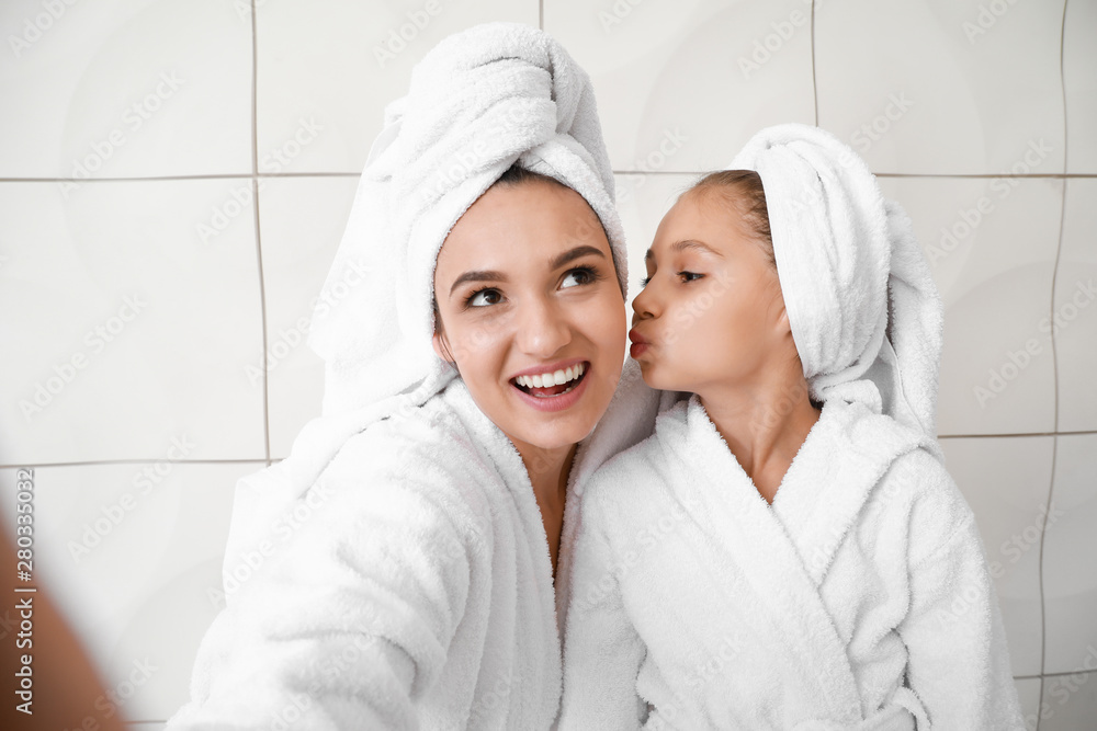 Mother and her little daughter in bathrobes taking selfie against white background