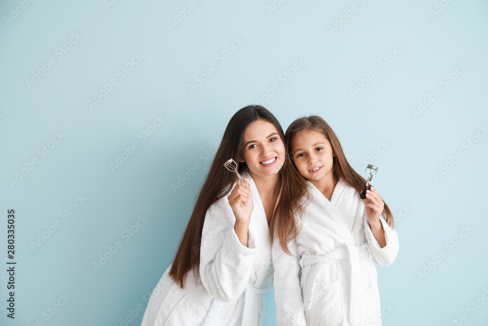 Mother and her little daughter in bathrobes and with eyelash curlers on color background