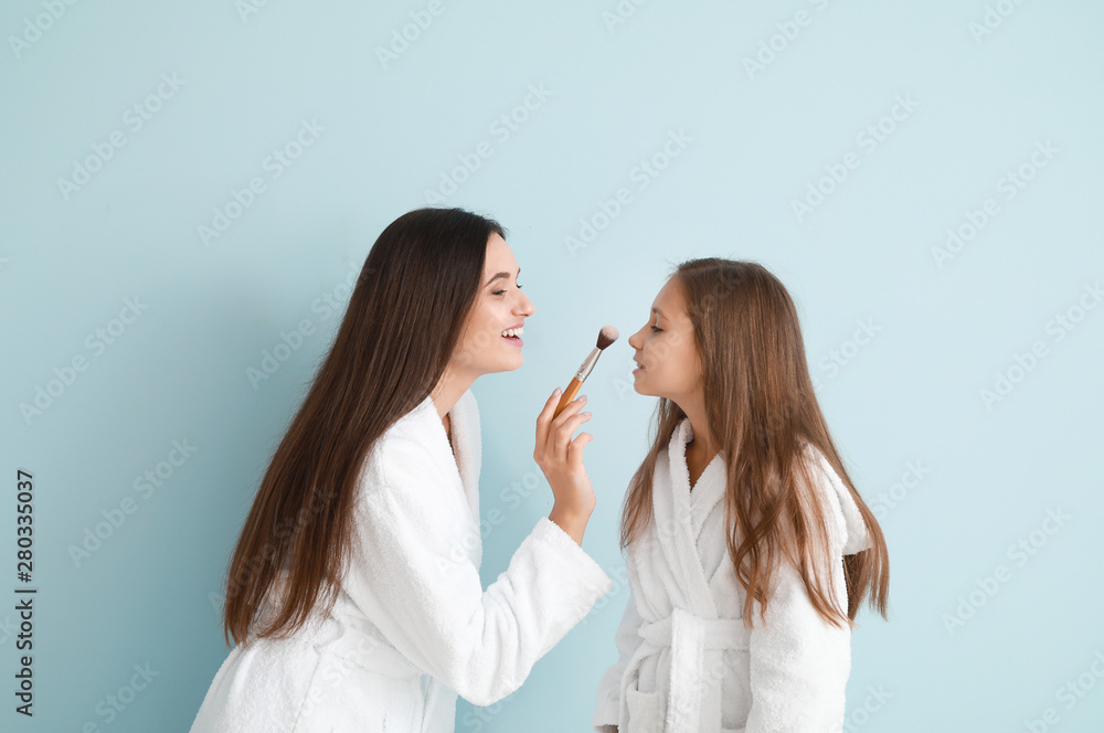 Mother and her little daughter in bathrobes applying makeup against color background