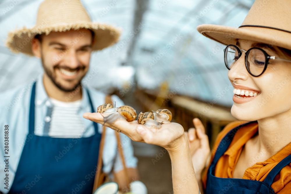 Two young beautifully dressed farmers examining snails growing process in the hothouse of the farm. 