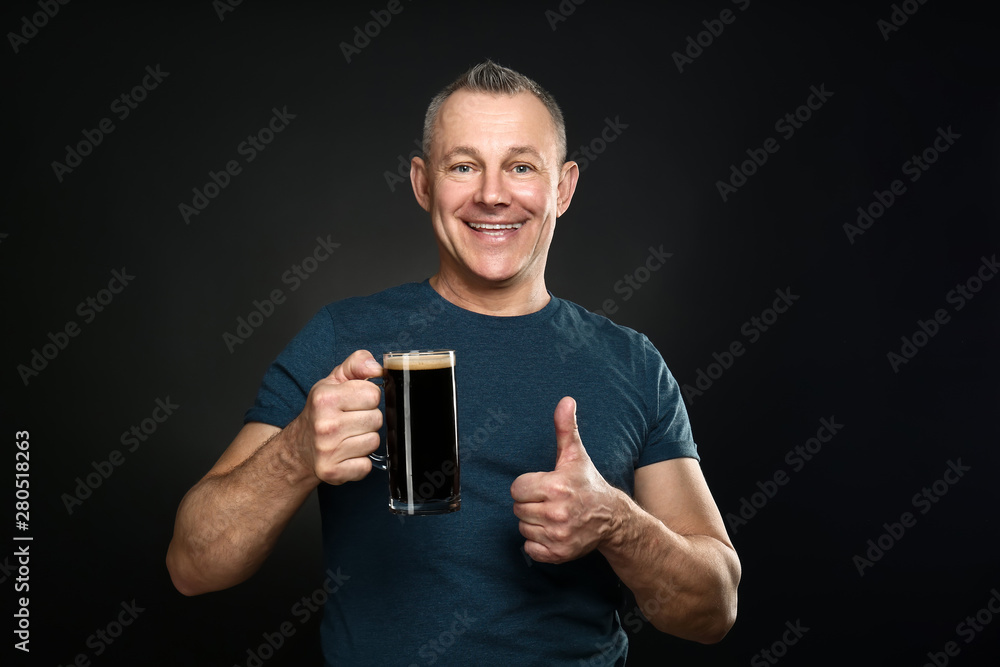 Happy middle-aged man with glass of beer showing thumb-up gesture on dark background