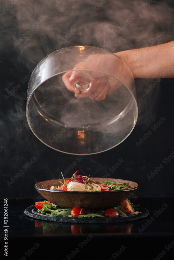 Tasty burnt caprese salad, cloche in hand, dark background.