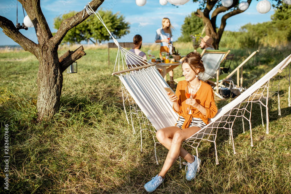 Young woman relaxing on the hammock, using smartphone during a lunch with friends in the garden on a