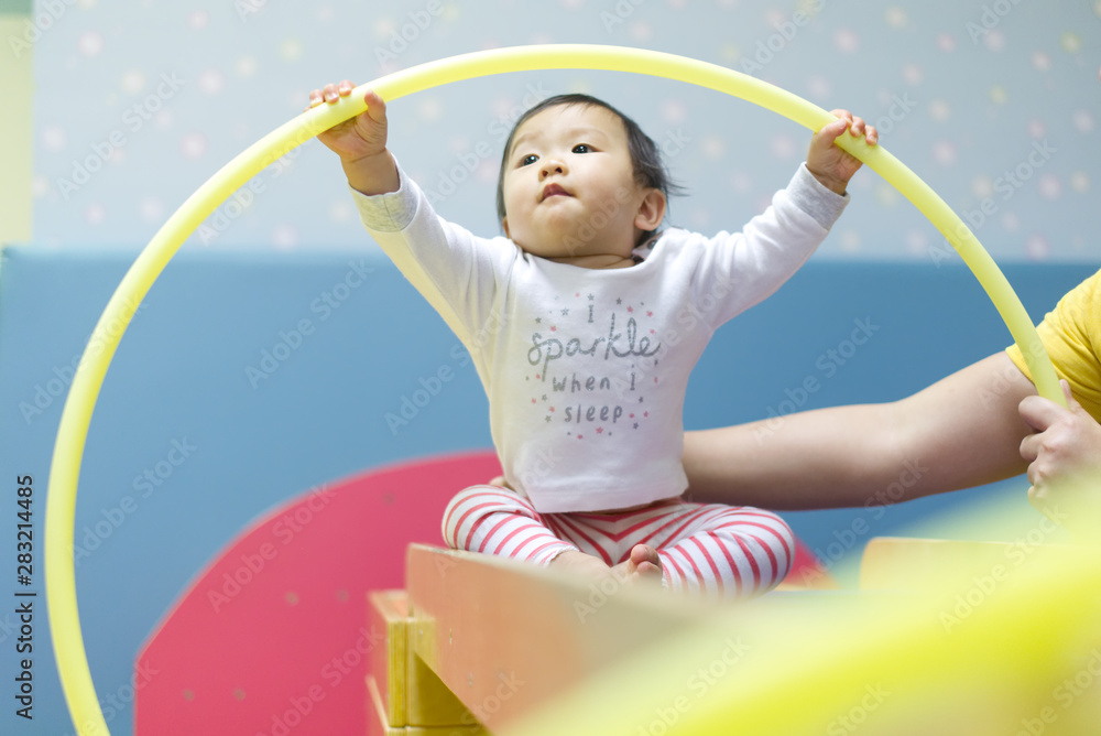 Young little Asian baby holding toy loop playing with preschool teacher in baby play gym. Kid enjoy 