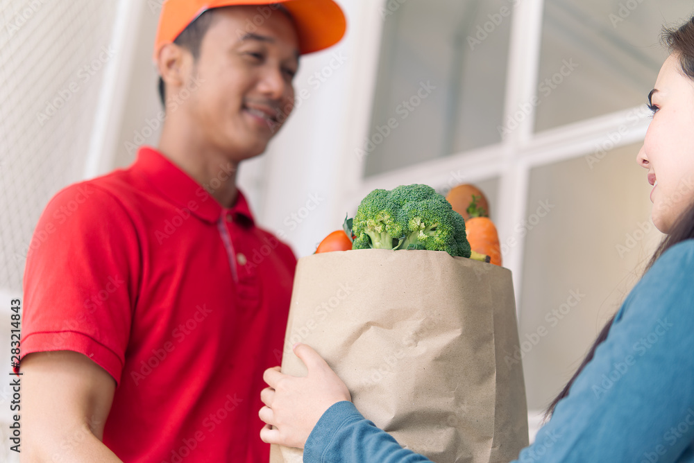 Asian delivery man in red color uniform handling bag of foods or vegetables to young beautiful femal