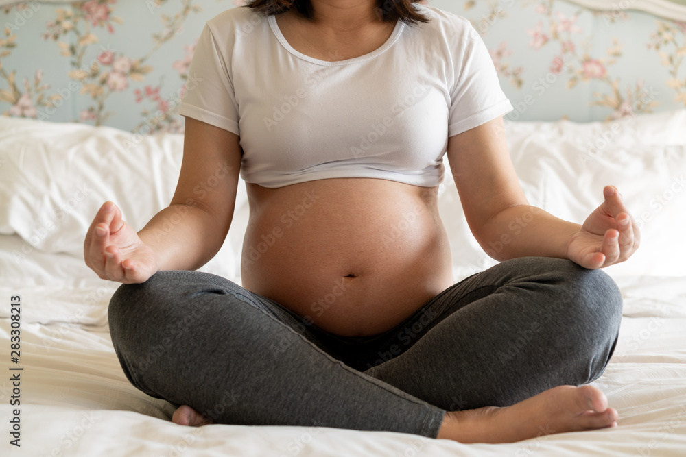 Pregnant woman doing yoga exercise on bed in bedroom at home while taking care of her child. The hap