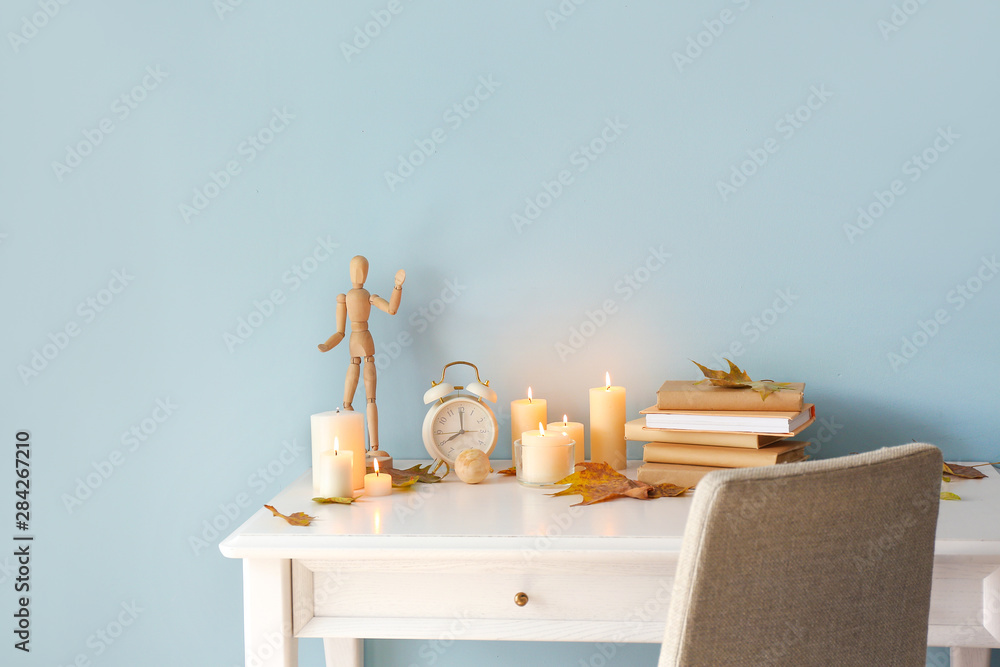 Table with beautiful burning candles, books, dry autumn leaves and decor near color wall