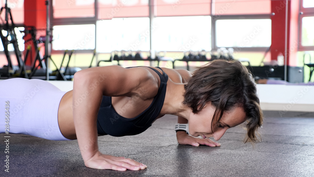 Sporty woman is doing set of reps press-ups from the floor in gym. She is spreading her arms wider a