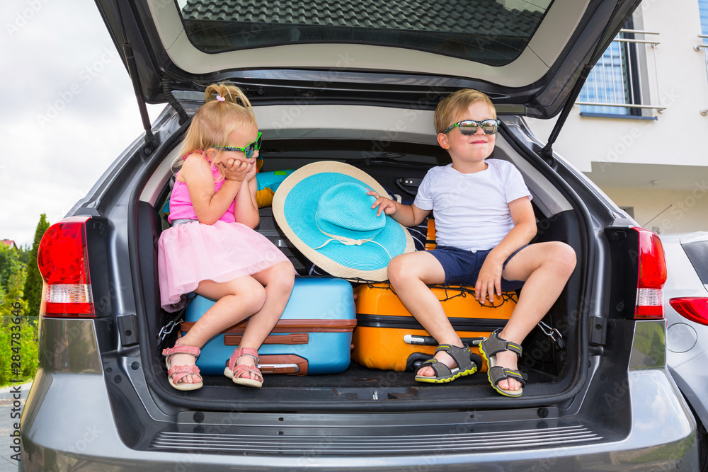 Twins boy and girl are sitting in the trunk of the car with luggage, ready to go for vacations.