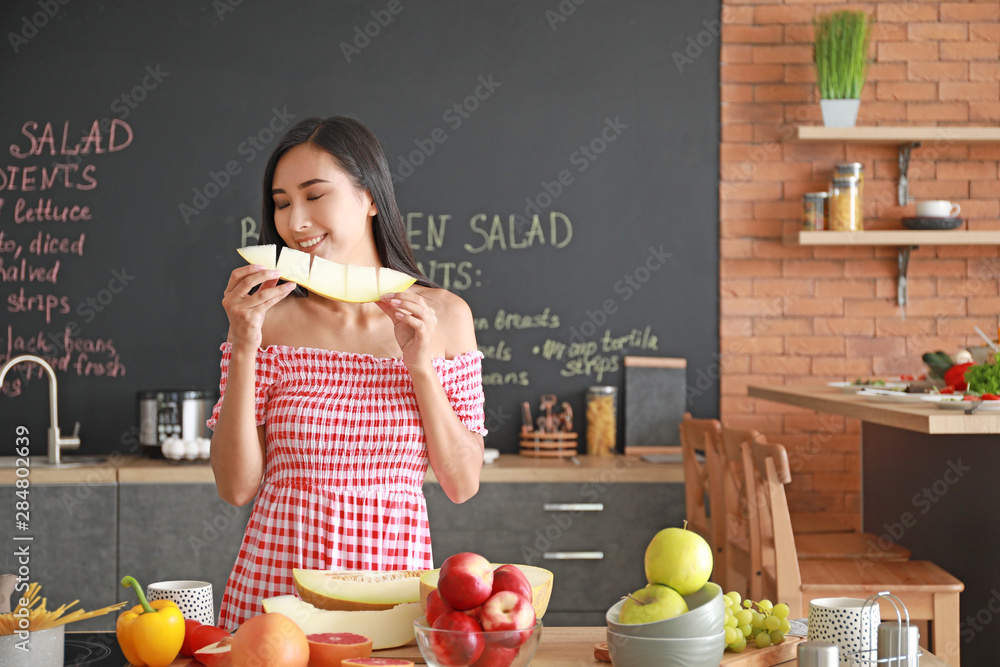 Beautiful Asian woman eating tasty melon in kitchen