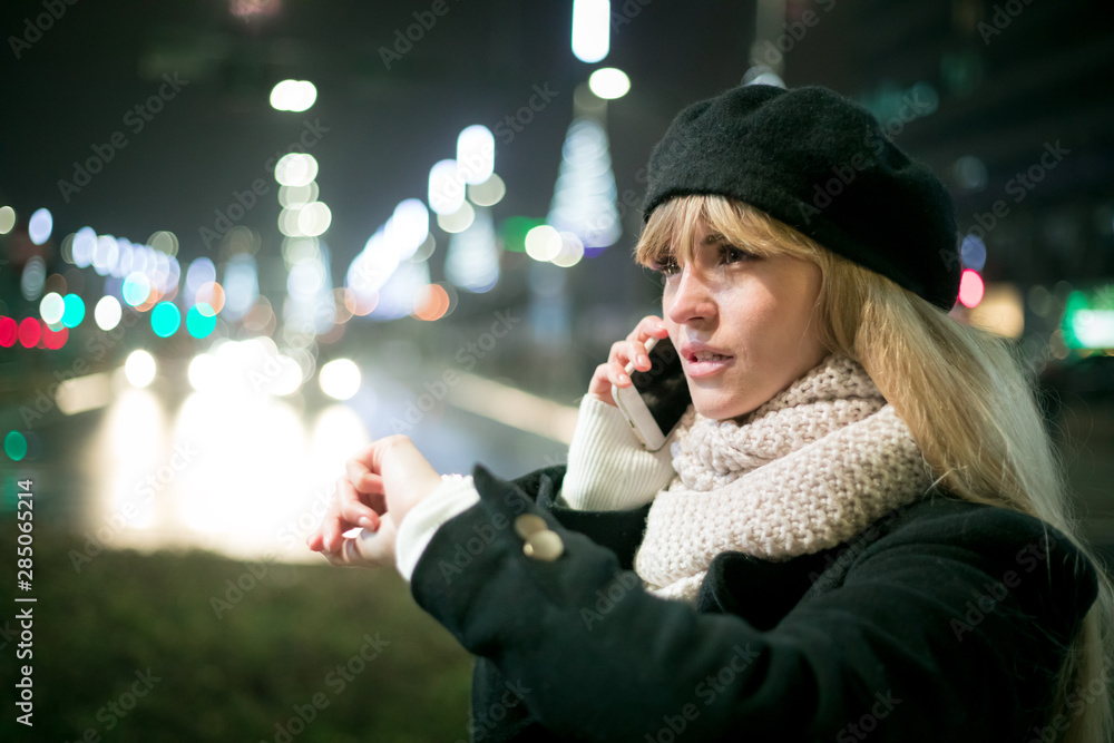 Pretty young stylish woman walking in the night city streets using modern smartpone, bokeh illuminat