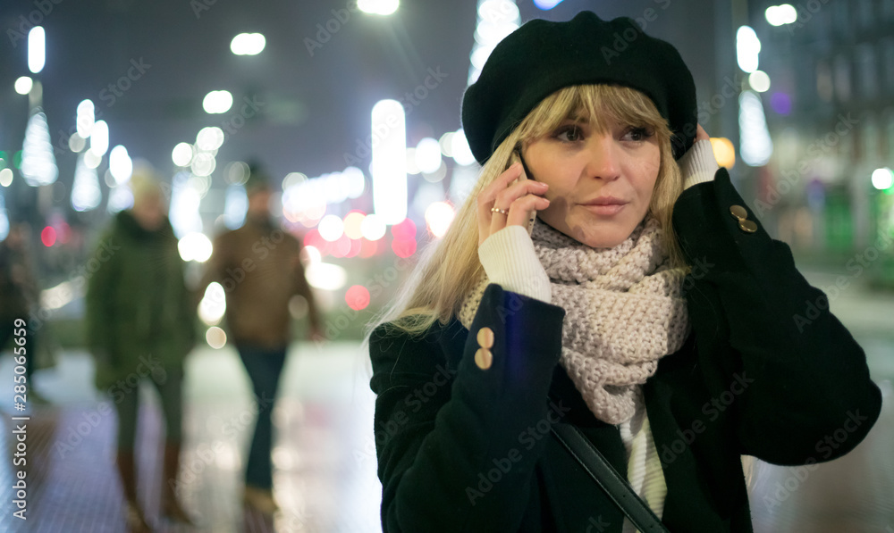 Pretty young stylish woman walking in the night city streets using modern smartpone, bokeh illuminat