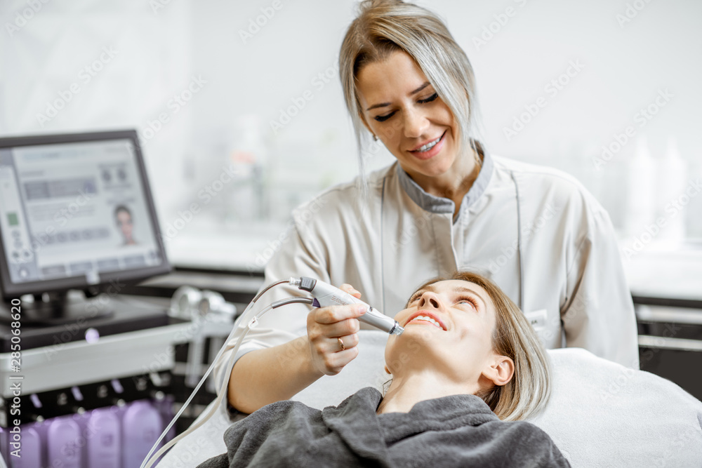 Cosmetologist making vacuum hydro peeling on the cheek area to a woman at the luxury beauty salon. C