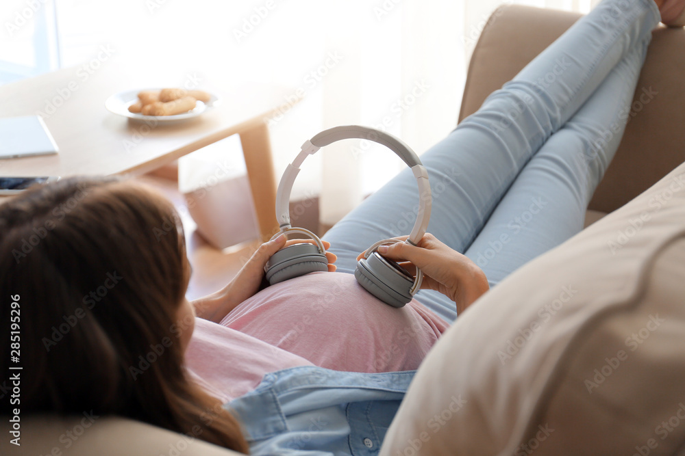 Beautiful pregnant woman putting headphones on her belly while lying on sofa