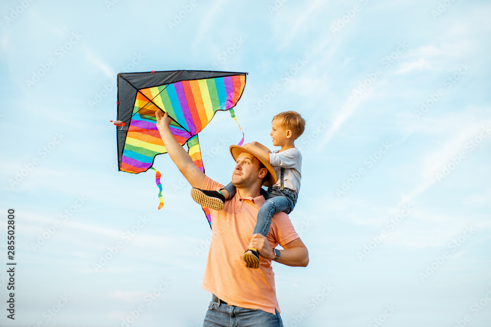 一位幸福的父亲和年幼的儿子的画像，肩上放着五颜六色的空中风筝，在蓝天上