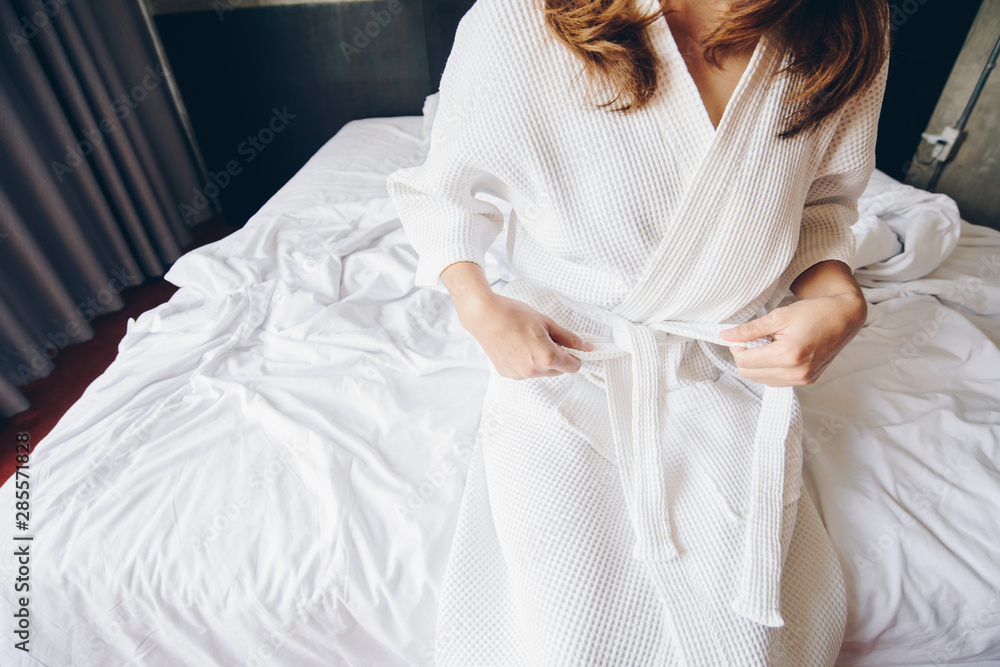 Cropped shot view of young woman sitting on the bed and tying up belt of white bathrobe before takin