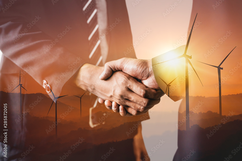 Double exposure graphic of business people handshake over wind turbine farm and green renewable ener