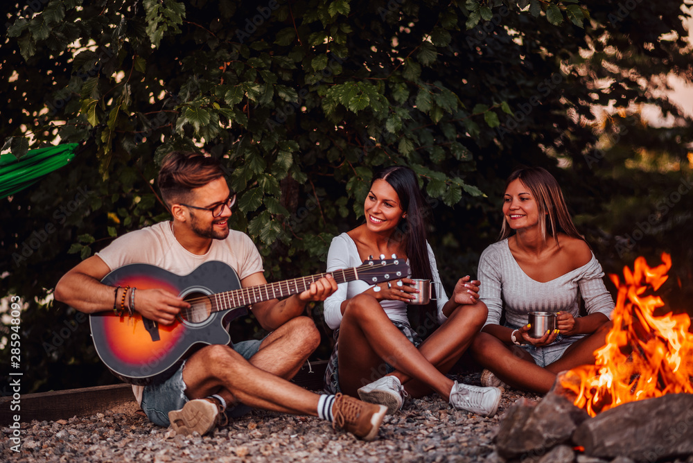 Group of friends camping. Sitting around camp fire, playing guitar.