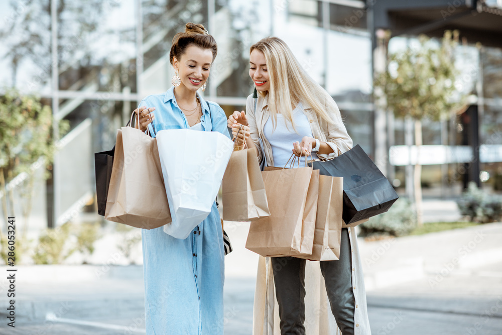 Two happy girlfriends feeling satisfied with purchases, standing together with shopping bags in fron