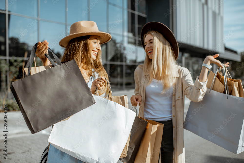 Two happy girlfriends walking with shopping bags in front of the shopping mall, feeling satisfied wi