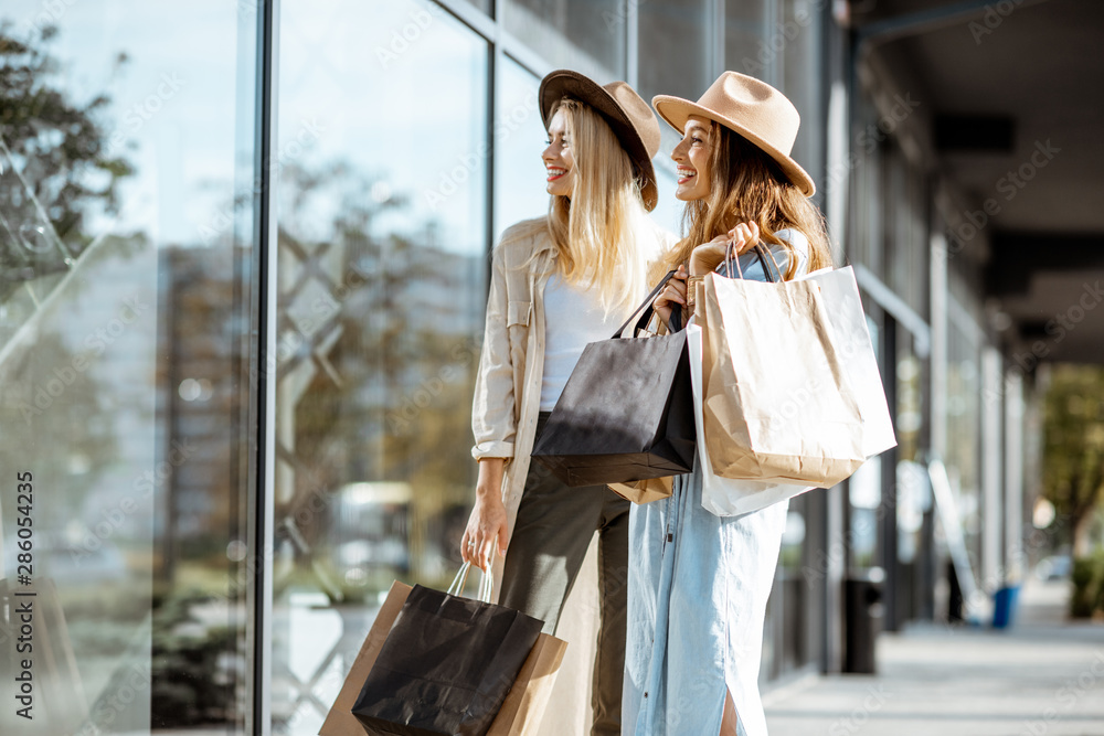 Two happy girlfriends looking on the shopwindow while standing with shopping bags near the mall