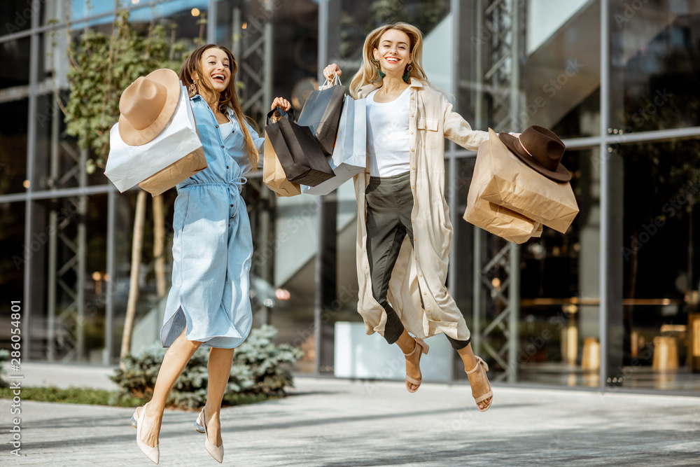 Two happy girlfriends jumpinh with shopping bags in front of the shopping mall outdoors, feeling exc