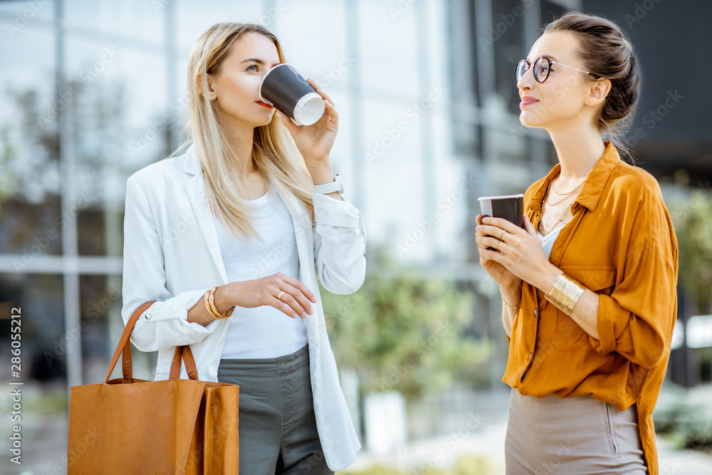 Two young businesswomen talking near the office building, having a small talk during the coffee brea