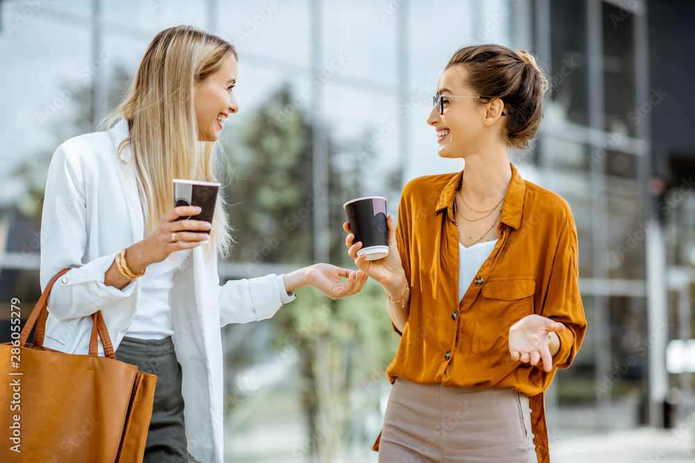 Two young businesswomen talking near the office building, having a small talk during the coffee brea