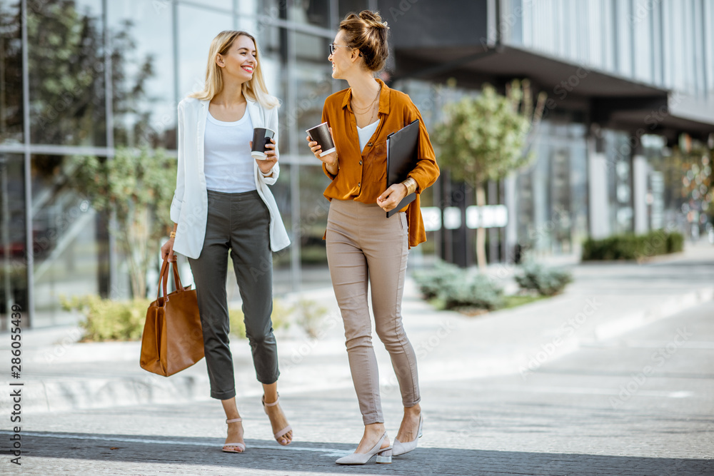 Full body portrait of a two young businesswomen walking with coffee cups near the modern office buil