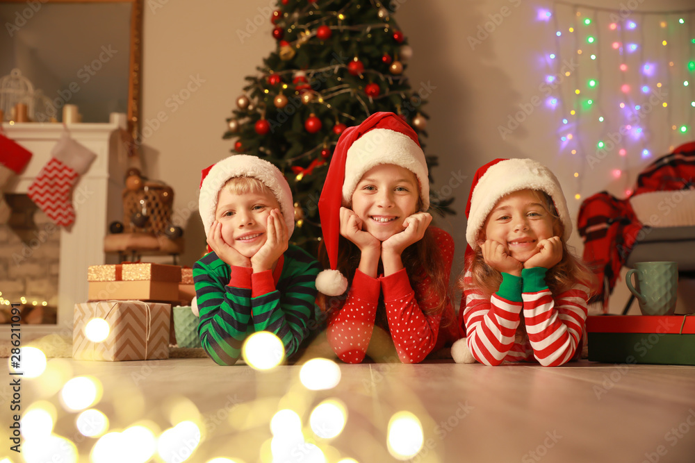 Cute little children in Santa Claus hats lying on floor on Christmas eve at home