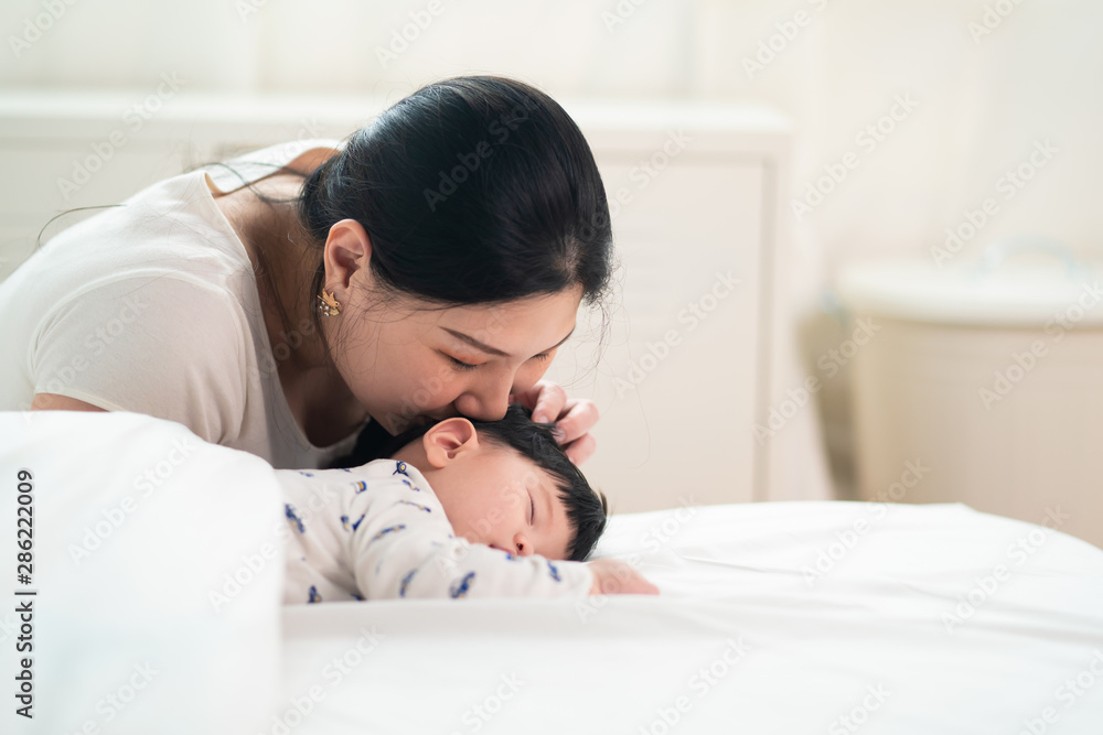 一位亚洲母亲在床上亲吻她可爱的熟睡新生儿的头。她微笑着伸出手