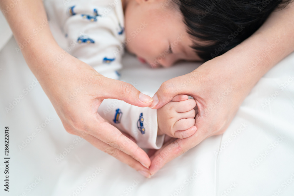 Top view of Asian mother is holding her cute sleeping newborn baby hand on bed. She making hands in 